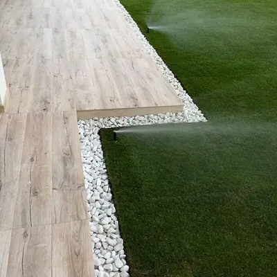 A modern backyard featuring a flawless lawn with an integrated pop-up sprinkler system in action. The lawn is edged with white pebbles, creating a sharp, clean border against the wood-look tile deck.