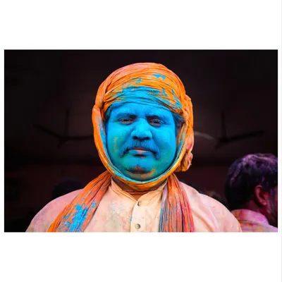 A man's face, covered in blue Holi powder, offers a moment of calm contemplation amidst the festival's chaos. His orange turban provides a striking color contrast.