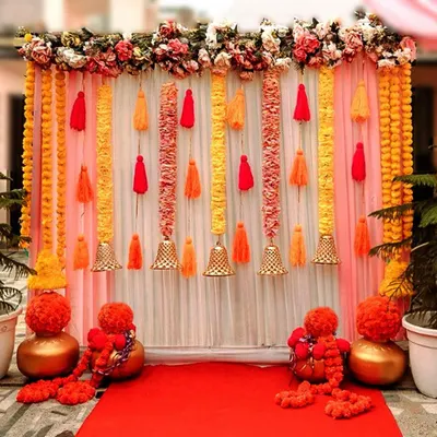 This bright and cheerful backdrop is a popular choice for Haldi and Mehendi functions. It features a combination of pink and white drapes, vertical garlands of marigolds, colorful tassels, and hanging bells, with decorative pots at the base.