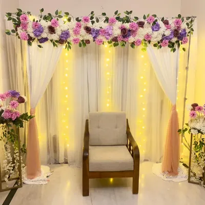 A simple and elegant backdrop for an at-home engagement or ceremony. The setup features a frame with white and pink drapes, a floral arrangement on top, and fairy lights for a warm glow.