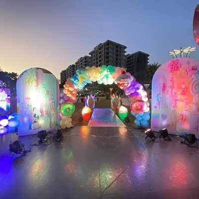 A spectacular outdoor Candyland party entrance at dusk, with large, illuminated backdrops, a rainbow balloon arch, and giant candy props.