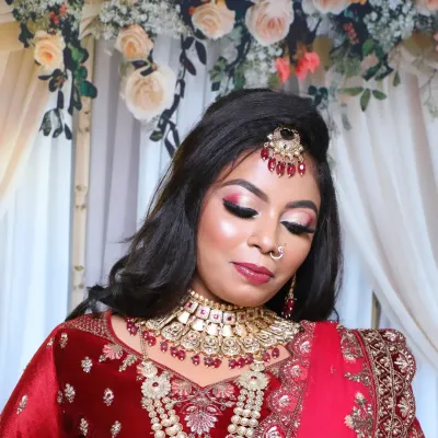 A close-up of a glamorous bridal look in a red lehenga. The makeup features dramatic red and pink eyeshadow, sharp contouring, and a full set of jewellery including a maang tikka and choker.
