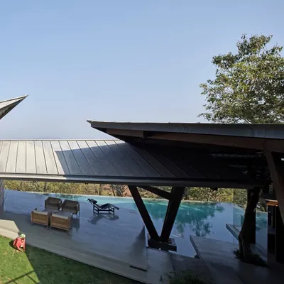 The Lateral Verandah House features a dramatic roofline that mirrors the stepping terrain. A bridge-like structure connects different parts of the home over the infinity pool.