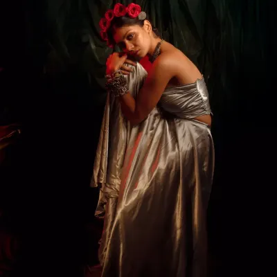A portrait from the 'INARA' series, styled to evoke a sense of romantic melancholy. The combination of a liquid silver top, heavy traditional jewelry, and a crown of red roses creates a powerful, goddess-like image.