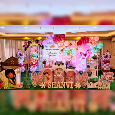 A beautiful and colorful butterfly theme for Shanvi's first birthday. The setup includes a lush green carpet, large butterfly wing cutout's, fairy character's, and a vibrant balloon backdrop.