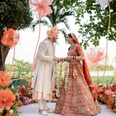 The couple stands at their beautiful outdoor mandap, surrounded by whimsical oversized paper flowers and a lush arrangement of burnt orange and peach florals.