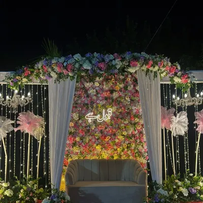 A breathtaking Nikah stage at night. The backdrop is a dense wall of colorful flowers with a 'Qubool Hai' neon sign, framed by a floral arch, white drapes, and sparkling string lights.