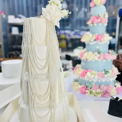 A beautiful comparison of two of our grand celebration cakes. On the left, a four-tier white cake with elegant pearl draping, and on the right, a five-tier pastel blue cake adorned with hundreds of handcrafted sugar flowers.