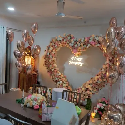 A beautiful heart-shaped floral and balloon arch set up for a romantic birthday dinner at home. The neon sign adds a modern touch, while the rose gold balloons create a warm, celebratory glow.