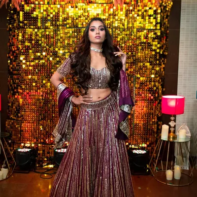 This is a full-on glamour look perfect for a sangeet or reception night. The makeup features a dramatic, colorful smokey eye and contoured cheeks, set against a glittering backdrop that matches her energy.