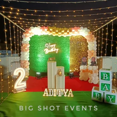 A complete setup for a second birthday, featuring a balloon arch, a green turf and gold sequin backdrop, marquee number '2', and personalized name and photo blocks.