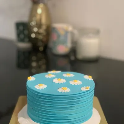 This daisy-themed bento cake is simple, elegant, and lovely. It features a textured blue buttercream base adorned with hand-piped daisies and a personalized name in green at the bottom.