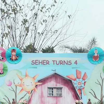 A close-up of the farm-themed birthday backdrop for "Seher Turns 4." The design features a cheerful sun, a pink barn, and a windmill, all in a cute, storybook art style.