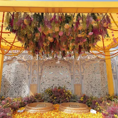 For this vibrant Haldi, the couple was seated amidst a garden of flowers. A canopy of hanging florals in shades of pink, yellow, and purple framed them against the venue's original thikri work wall, creating a stunning visual.