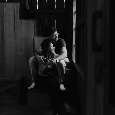 A quiet, contemplative moment captured in black and white. The couple sits by a window, bathed in soft, natural light, creating a timeless and soulful portrait.