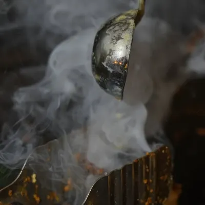 This artistic shot captures a chef adding a tempering of hot spices, with a billow of aromatic smoke. It represents the culinary craft and the authentic cooking techniques we use to create flavorful Indian dishes.