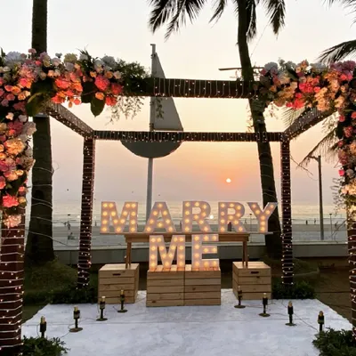 The perfect proposal setting at sunset. A beautiful floral arch frames the sea view, with 'MARRY ME' marquee lights ready for the big question.