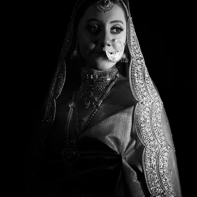 A dramatic black and white bridal portrait using high-contrast lighting to create a powerful and mysterious image. The focus is entirely on her expression and the intricate details of her attire.