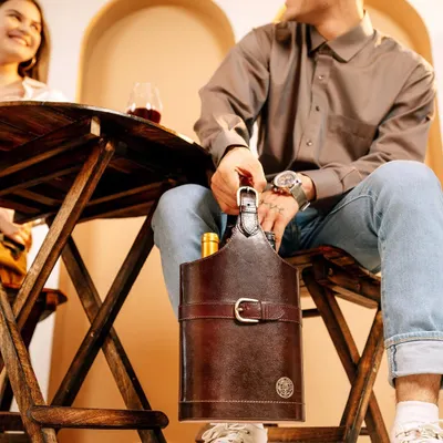 A lifestyle scene for Verman Atelier's leather wine carrier. The shot captures a candid moment between a couple, with the product looking stylish and functional in a social, sophisticated setting.