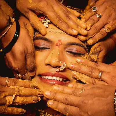 A close up of the bride during her Haldi ceremony. Surrounded by the hands of her loved ones applying turmeric paste, her smile shows pure happiness.