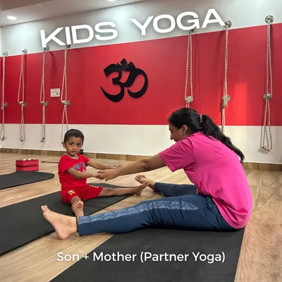 A mother and son practicing a seated forward bend together during a partner yoga class. These sessions are a wonderful opportunity for parents to connect with their children on a different level, fostering teamwork and mutual support in a fun, healthy environment.