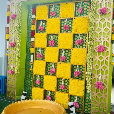 A vibrant Haldi backdrop with a checkerboard pattern of yellow marigolds. The setup includes a traditional brass vessel (urli) for the ceremony, framed by decorative panels.