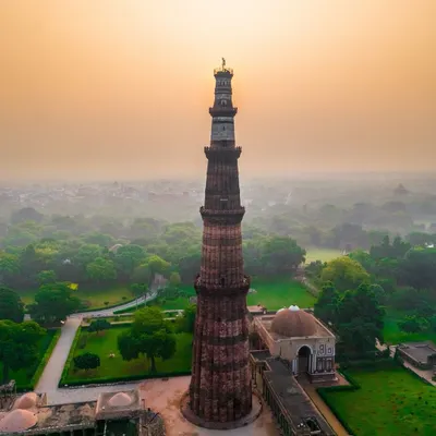 The Qutub Minar in Delhi stands tall against a hazy sunrise. This aerial shot captures the monument and its surrounding lush greenery from a unique, elevated perspective.