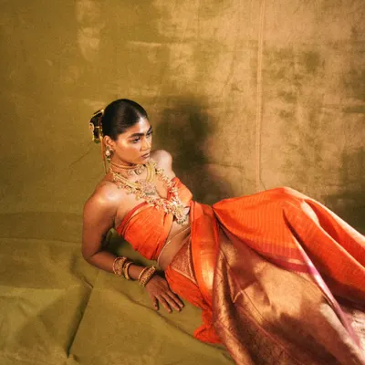 A full-length view of the 'Swaṟṇavalli' concept, where the rich orange of the silk sari is complemented by warm, bronzed skin and traditional gold jewellery. The makeup was designed to feel both regal and ethereal.