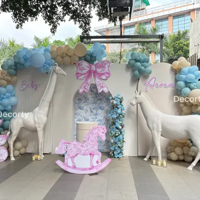 A grand Dior-inspired baby shower backdrop with a soft blue and pink color scheme. The setup features life-sized animal props like a giraffe and unicorn, a rocking horse, and a vintage-style pram cutout for a touch of elegance.