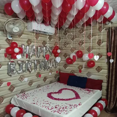 A classic romantic birthday room decoration with red and white balloons on the ceiling and floor, "Happy Birthday" foil letters, and a heart made of rose petals on the bed.