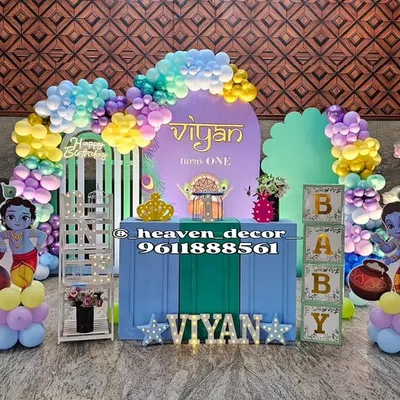 A vibrant and colorful Little Krishna theme for a first birthday. The setup features multiple Krishna cutouts, a rainbow balloon arch, and decorative elements like lotus flower props and "BABY" blocks.