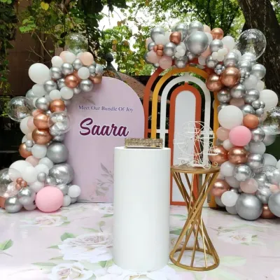 A close-up of a modern and chic naming ceremony backdrop for "Saara". The decor features a pastel color palette with rose gold, silver, and pink balloons, a custom arched backdrop, and elegant pedestals.