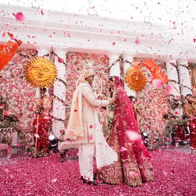 A perfect Varmala moment captured amidst a shower of pink flower petals. The couple is framed by dancers with sun props and a stunning floral wall, creating a scene of pure romance and grandeur.