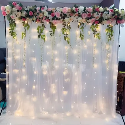 A beautiful floral backdrop for an indoor event. The top of the frame is covered in pink and white artificial flowers with hanging green vines and a fairy light curtain.