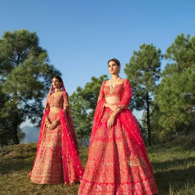 A full view of two bridal lehengas for a Frontier Raas campaign. The styling showcases two distinct but complementary red ensembles against a lush, green mountain landscape, highlighting the grandeur of the outfits in a natural setting.