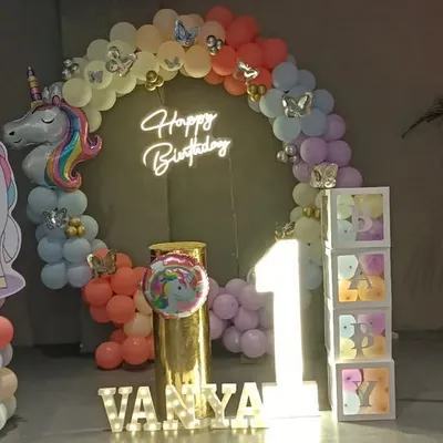 A magical unicorn-themed setup for Vanya's first birthday. It features a circular backdrop with a neon sign, unicorn cutouts, a light-up number '1', and a pastel rainbow balloon arch.