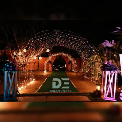 A magical entrance tunnel created with arches of fairy lights, leading guests into a wedding event over a green carpet walkway.