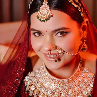A stunning close-up of the bride, her face illuminated by a warm red light. This portrait beautifully captures her flawless makeup, intricate nose ring, and the heavy diamond and emerald necklace.