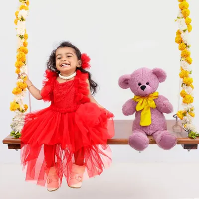 A picture of pure joy. This happy toddler is dressed in a bright red dress, sitting on a flower-adorned swing with her teddy bear companion for a fun and whimsical portrait.