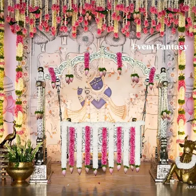 A beautiful cradle ceremony setup inspired by traditional art. The centerpiece is a floral-decorated swing set against a Pichwai-style backdrop featuring Lord Krishna, surrounded by brass props and flower arrangements.