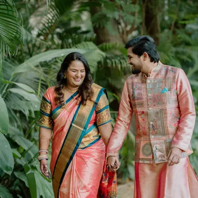 A candid close-up of a couple in beautiful traditional outfits, holding hands and smiling at each other while walking through a park. Their genuine laughter is the focus of this engagement photo.