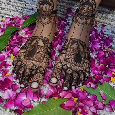 A stunning bridal leg design featuring a peacock portrait within a frame, surrounded by intricate patterns and a lotus anklet band. The design is beautifully presented on a bed of flower petals.