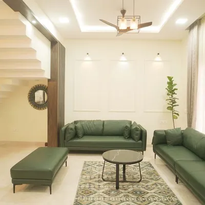 This spacious villa living room is designed for serene comfort, featuring plush green leather sofas, minimalist wall paneling, and large windows that fill the space with natural light. The open layout connects seamlessly to the staircase, creating a sense of grandeur and flow.
