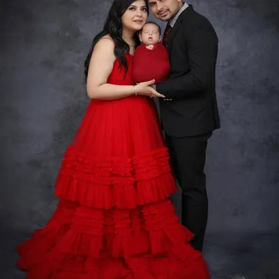 A formal family portrait featuring parents dressed elegantly with their newborn swaddled in a matching deep red wrap. The mother's tiered red gown adds a touch of drama and elegance.