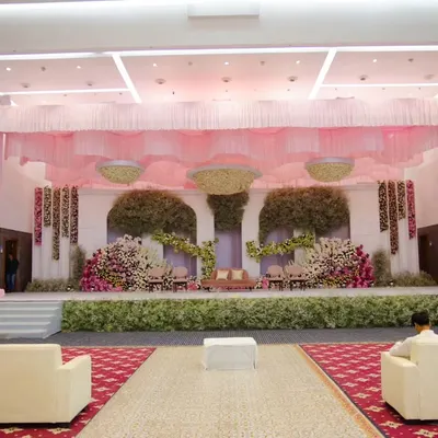A full view of a grand ballroom setup featuring a soft pink and green color palette. The stage is framed with draped fabric and lush florals, creating an elegant backdrop for a large-scale reception or corporate gala.