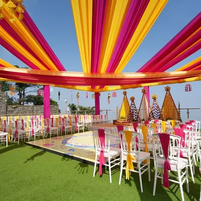 An expansive view of a stunning rooftop Haldi ceremony. We used vibrant yellow and pink drapes to create a festive canopy against the blue sky, with a beautifully decorated stage and seating for all the guests.