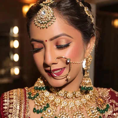 A classic bridal portrait showcasing a flawless HD base, warm smokey eyes, and a rich red lip. The look is completed with traditional gold and green kundan jewelry, perfect for a timeless wedding ceremony.
