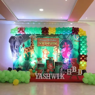 A fun jungle safari theme for Yashwik's birthday. This stage decoration includes a vibrant green backdrop, cutouts of various jungle animals like a giraffe and elephant, and a matching green balloon frame.