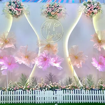 This photo booth features a backdrop of large, handcrafted paper flowers in pastel shades, accented with a custom "All for Love" neon sign. The white picket fence adds a charming, garden-like feel.