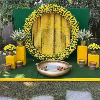 A classic Haldi backdrop for outdoor functions. This setup features a large circular frame filled with strings of yellow marigolds, a green panel background, and a traditional brass urli in the center for the ceremony.